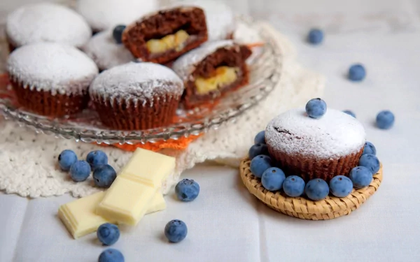 A beautifully arranged display of cupcakes, some filled with cream and dusted with powdered sugar, surrounded by fresh blueberries and white chocolate, serving as a vibrant HD desktop wallpaper.