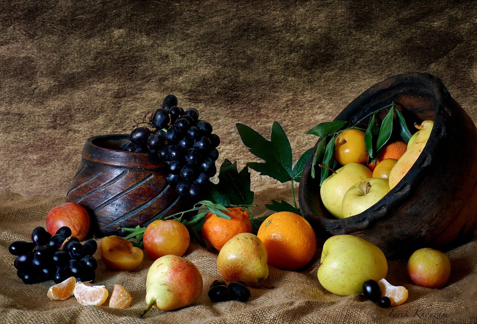 A vibrant arrangement of fruits, including grapes, oranges, and pears, displayed with an antique pot on a textured background, creating an inviting HD desktop wallpaper.