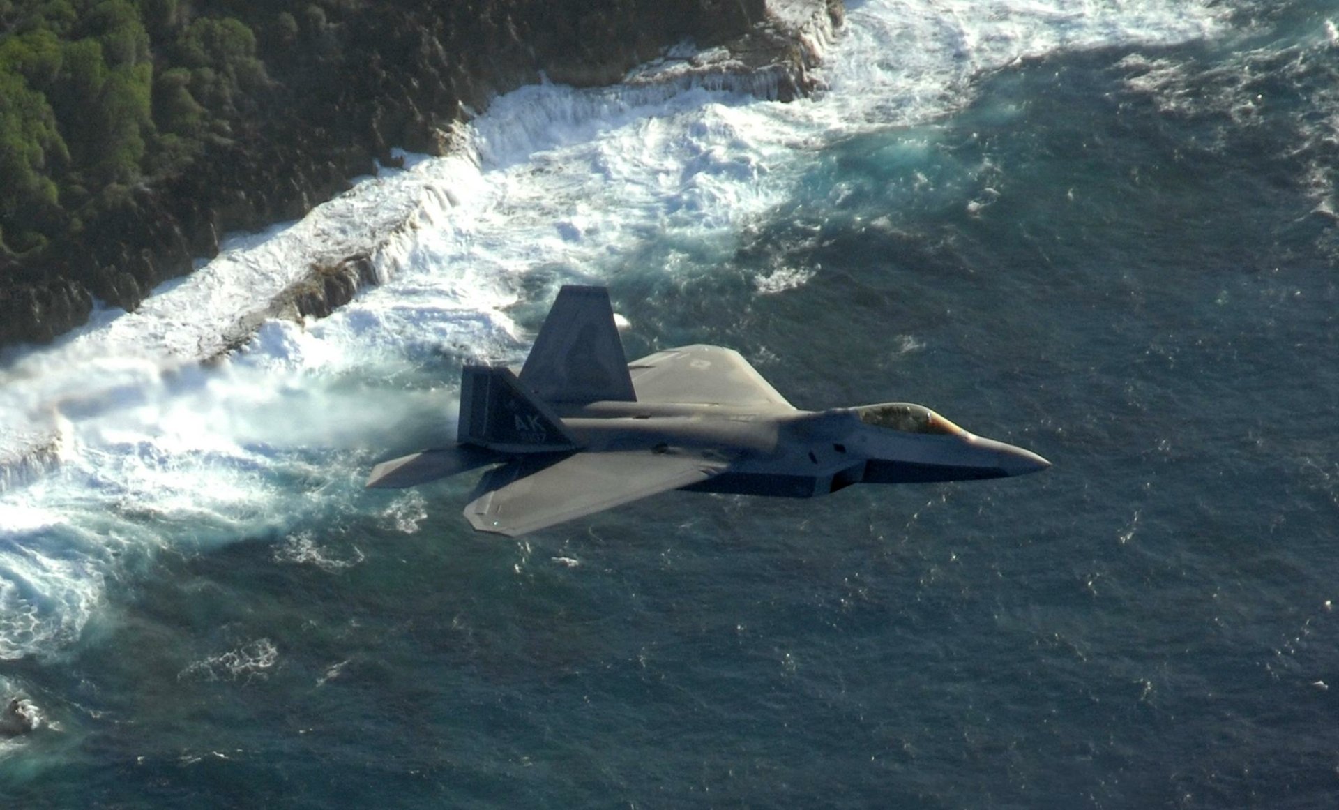 A Lockheed Martin F-22 Raptor military jet flying over a rugged coastline with waves crashing against the rocks, captured in high definition for a desktop wallpaper.