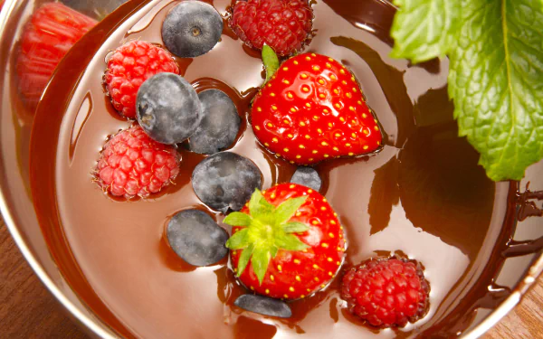 A close-up of fresh strawberries, raspberries, and blueberries dipped in rich chocolate, surrounded by vibrant green mint leaves, creating an enticing dessert scene for HD wallpaper.