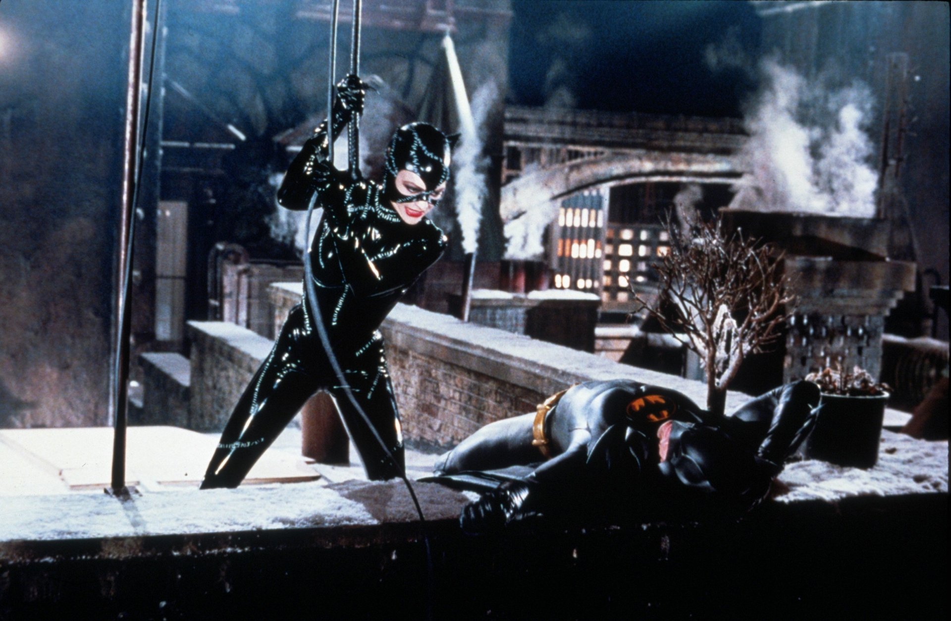 Batman Returns 2K Quad HD PC desktop wallpaper: Catwoman in glossy black suit on a snowy rooftop, poised over a fallen Batman as gothic city smoke and lights glow behind them.