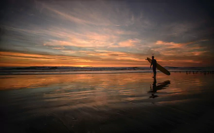 A silhouette of a surfer carrying a surfboard walks along the beach at sunset, reflecting the vibrant colors of the sky on the wet sand, capturing the essence of surfing and sports.