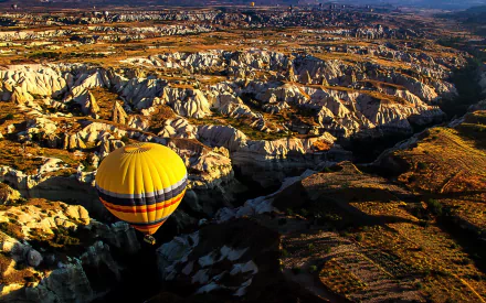 HD desktop wallpaper showing a colorful hot air balloon flying over a rugged, sunlit canyon landscape with deep shadows and detailed rock formations.