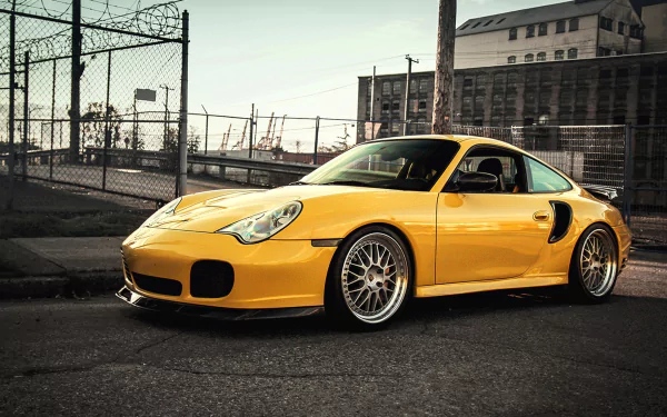 Yellow Porsche sports coupe parked on an urban street beside a chain-link fence and industrial buildings — HD PC desktop wallpaper and background.