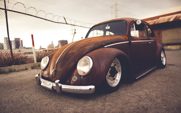 A rust-colored Volkswagen Beetle parked outdoors, featured as an HD PC desktop wallpaper and background with an industrial fence and buildings in the background.