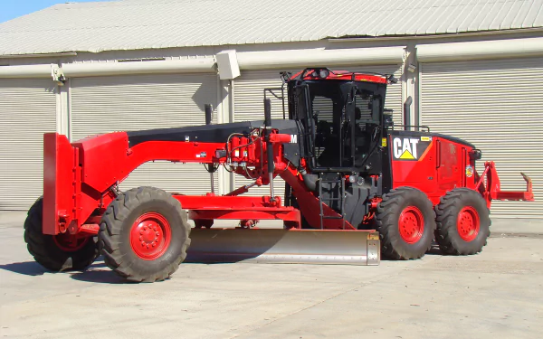 HD desktop wallpaper showcasing a bright red Caterpillar grader vehicle parked on a concrete surface in front of a large industrial building.