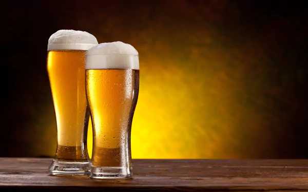 Two glasses of golden beer with frothy tops sit on a wooden surface, set against a warm, glowing background. This HD desktop wallpaper captures the essence of food and drink.