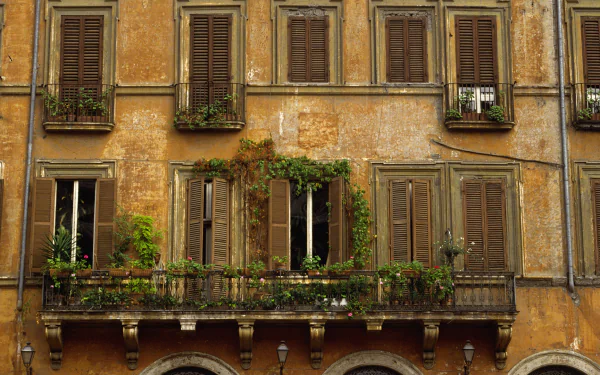 HD desktop wallpaper of traditional Roman architecture featuring a weathered building facade with wooden shutters and a balcony adorned with green plants in Italy.