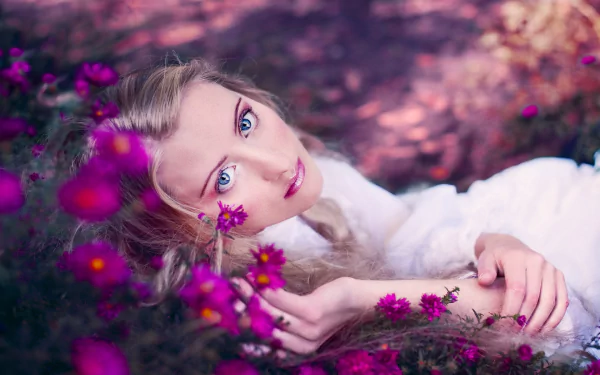 A beautiful woman with captivating blue eyes lies among vibrant pink flowers in this HD desktop wallpaper and background.
