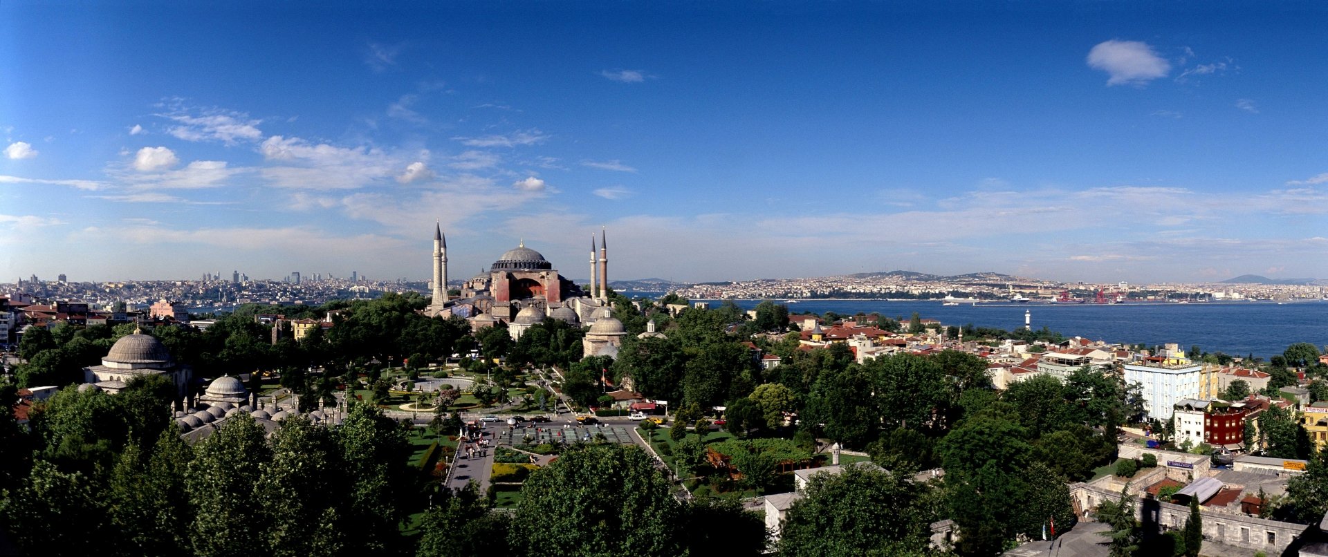 A breathtaking HD wallpaper featuring Hagia Sophia, surrounded by lush greenery and a vibrant cityscape, under a clear blue sky, embodying its rich religious and historical significance.