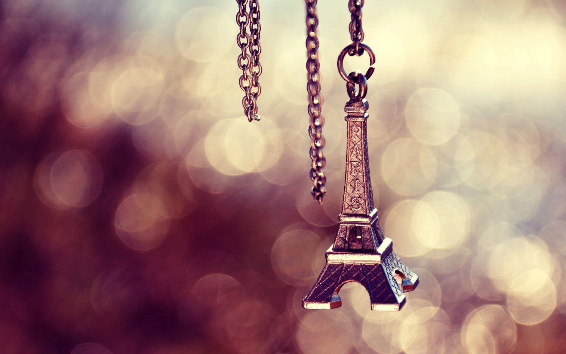A close-up of a miniature Eiffel Tower pendant hanging from a chain, set against a blurred, colorful background, creating a captivating desktop wallpaper.
