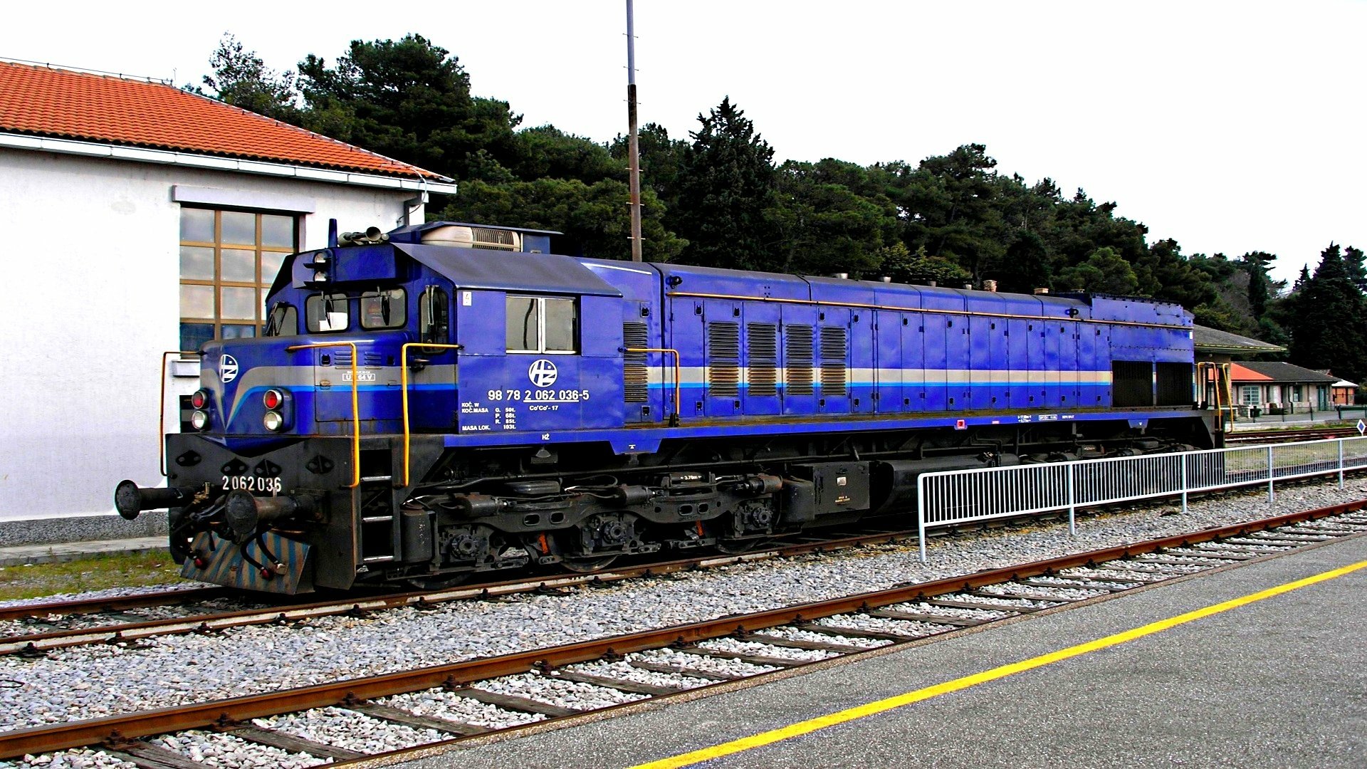 HD desktop wallpaper featuring a blue train locomotive stationed on railway tracks beside a platform with trees and a building in the background.