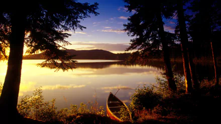 HD wallpaper of a serene lake at sunset, framed by silhouetted trees, with a canoe resting by the shore.