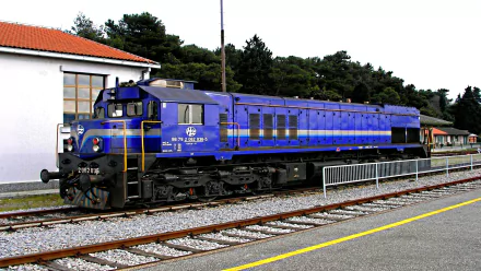 HD desktop wallpaper featuring a blue train locomotive stationed on railway tracks beside a platform with trees and a building in the background.