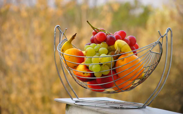 A vibrant assortment of fruits in a wire basket, set against a blurred autumn backdrop, captured in stunning 4K Ultra HD, serving as a colorful desktop wallpaper.
