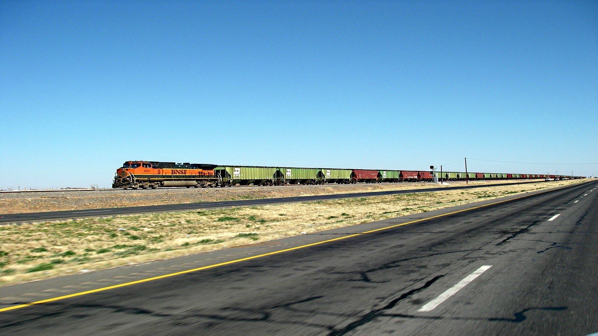 HD desktop wallpaper showcasing a long freight train traveling on parallel tracks through a flat, open landscape under a clear blue sky.