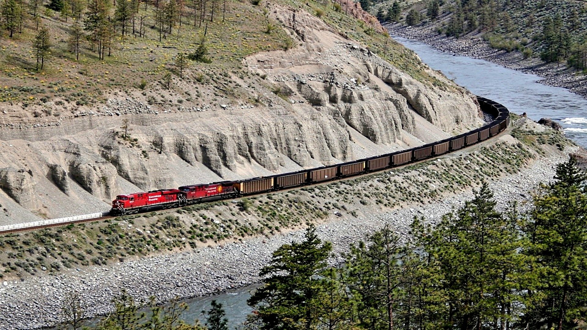 A train travels along a winding track beside a river, surrounded by lush greenery and rugged terrain, creating a captivating scene for a high-definition desktop wallpaper.