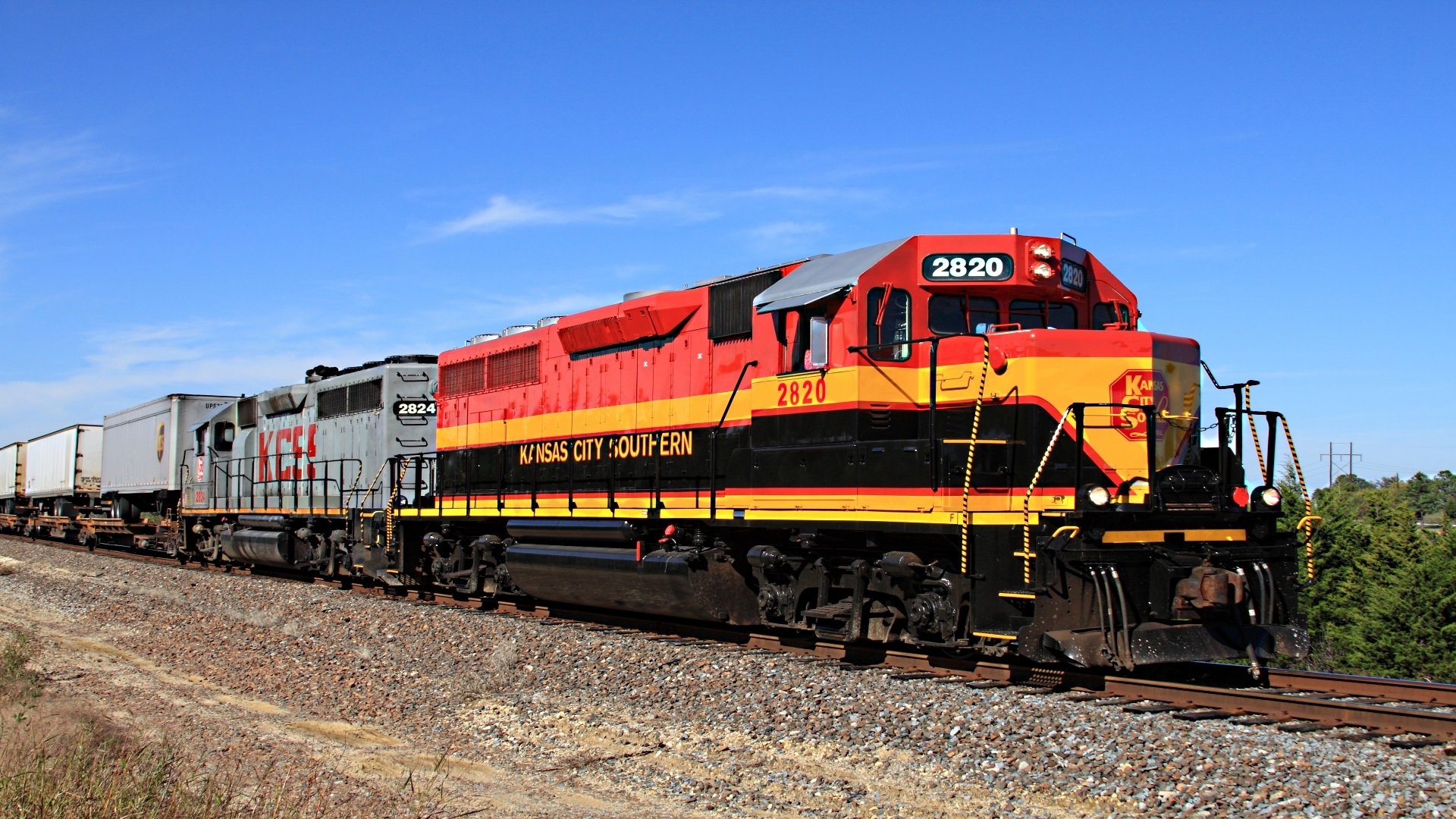 HD desktop wallpaper featuring a vibrant Kansas City Southern train engine traveling on railroad tracks under a clear blue sky.