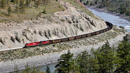 A train travels along a winding track beside a river, surrounded by lush greenery and rugged terrain, creating a captivating scene for a high-definition desktop wallpaper.