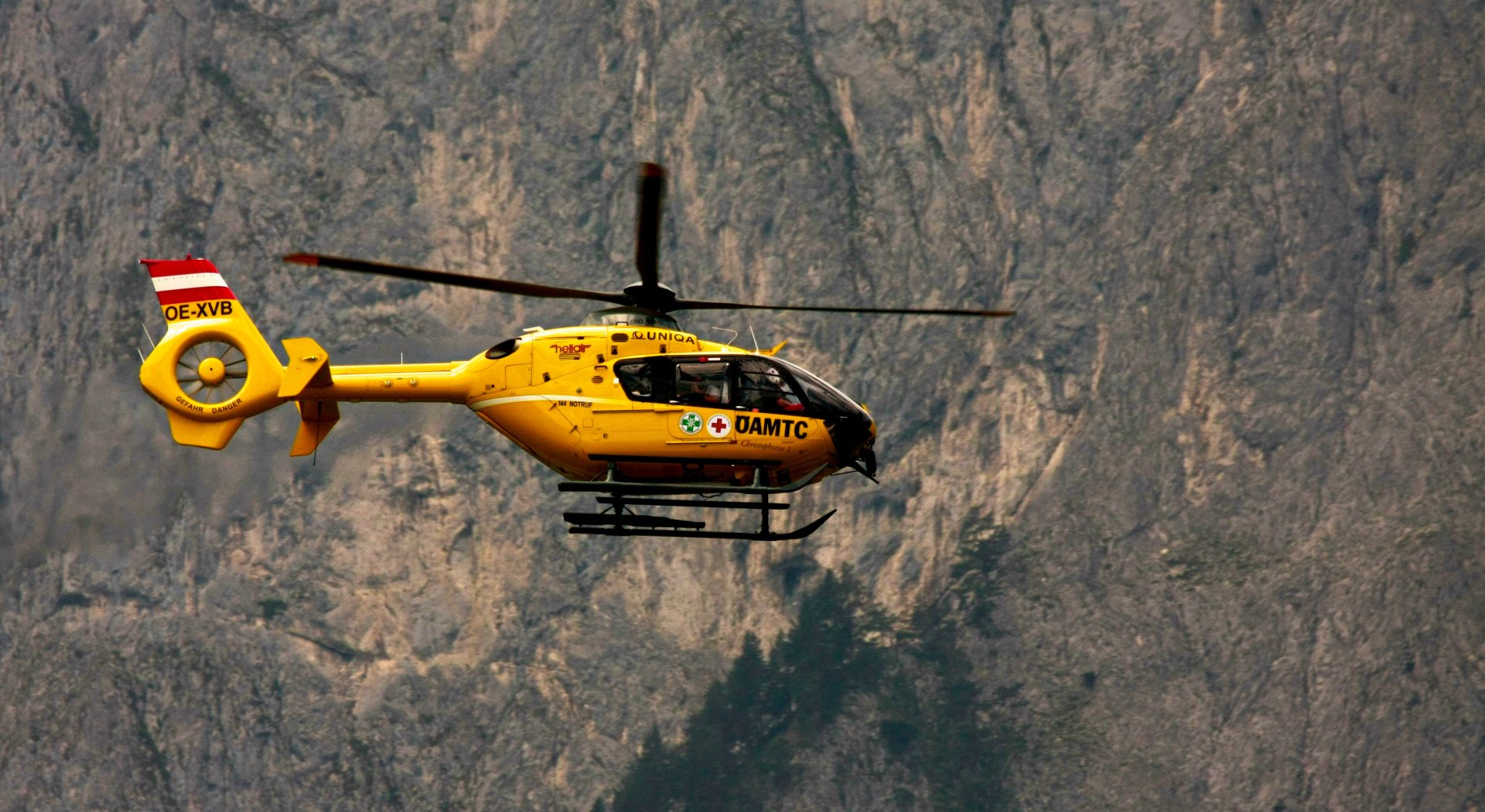 Yellow Eurocopter EC135 helicopter flying against a rugged mountain backdrop, captured in a high-definition desktop wallpaper image.