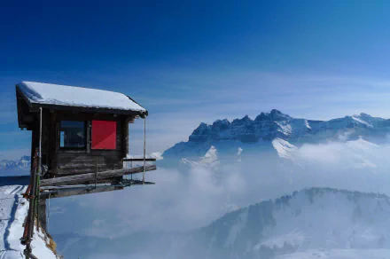 A wooden cabin perched on a snowy mountain cliff surrounded by ice and cold, overlooking misty snow-covered peaks under a clear blue sky.