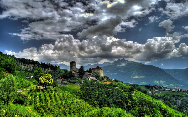 Panoramic view of a South Tyrol village in Italy featuring a historic castle nestled among green vineyards and mountains under a dramatic cloudy sky.