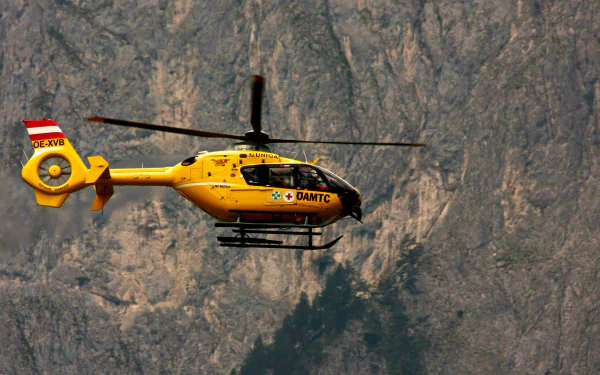 Yellow Eurocopter EC135 helicopter flying against a rugged mountain backdrop, captured in a high-definition desktop wallpaper image.