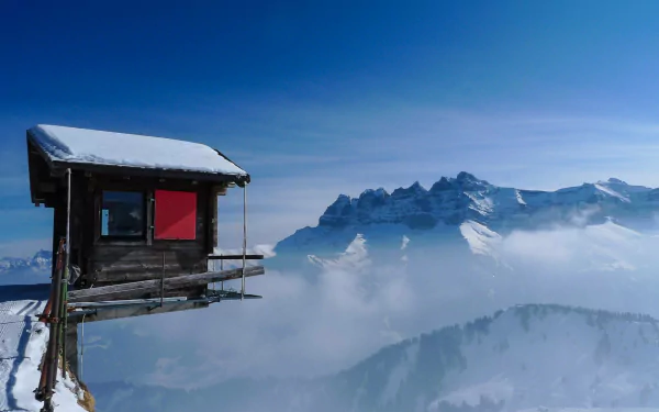 A wooden cabin perched on a snowy mountain cliff surrounded by ice and cold, overlooking misty snow-covered peaks under a clear blue sky.