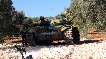 HD PC desktop wallpaper of a camouflaged T-55 tank in an olive grove, tracks in churned earth and personnel beside it under a clear blue sky — military background.