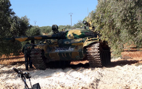 HD PC desktop wallpaper of a camouflaged T-55 tank in an olive grove, tracks in churned earth and personnel beside it under a clear blue sky — military background.