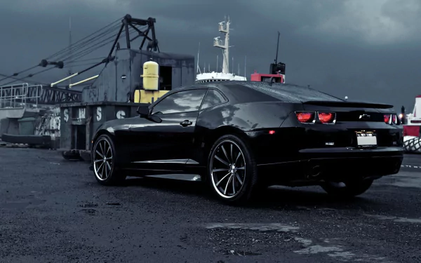 HD PC desktop wallpaper featuring a sleek black Chevrolet Camaro parked on an industrial waterfront under a cloudy sky.