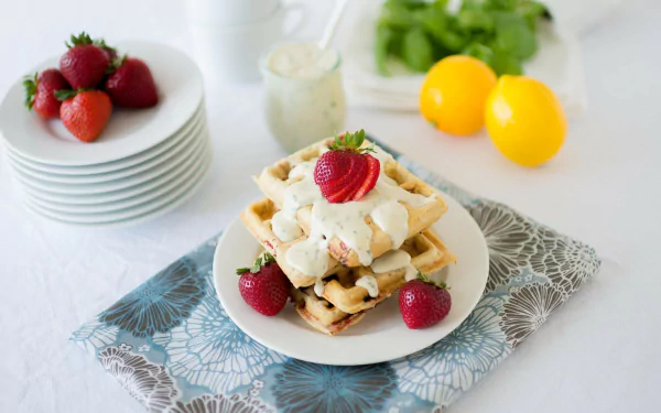 HD desktop wallpaper featuring a stack of waffles topped with whipped cream and strawberries, with fresh strawberries, lemons, and a glass jar in the soft-focused background.