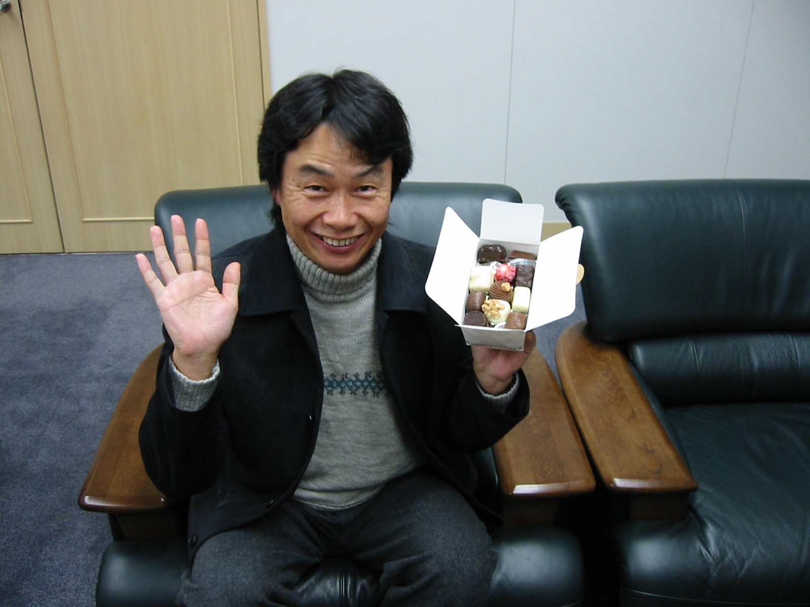 A man, Shigeru Miyamoto, smiles and waves while holding a box of assorted sweets, seated comfortably on a couch. This image serves as a vibrant HD desktop wallpaper and background.