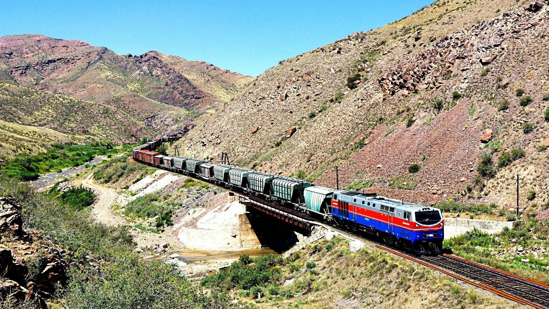 A vibrant train navigates a scenic landscape, crossing a bridge against a backdrop of rugged mountains. This HD image makes an impressive desktop wallpaper or background.