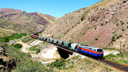A vibrant train navigates a scenic landscape, crossing a bridge against a backdrop of rugged mountains. This HD image makes an impressive desktop wallpaper or background.
