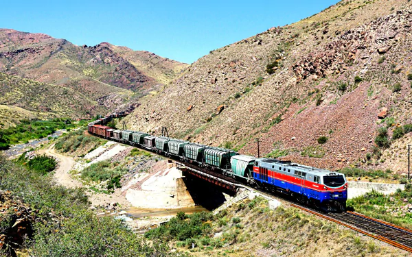 A vibrant train navigates a scenic landscape, crossing a bridge against a backdrop of rugged mountains. This HD image makes an impressive desktop wallpaper or background.