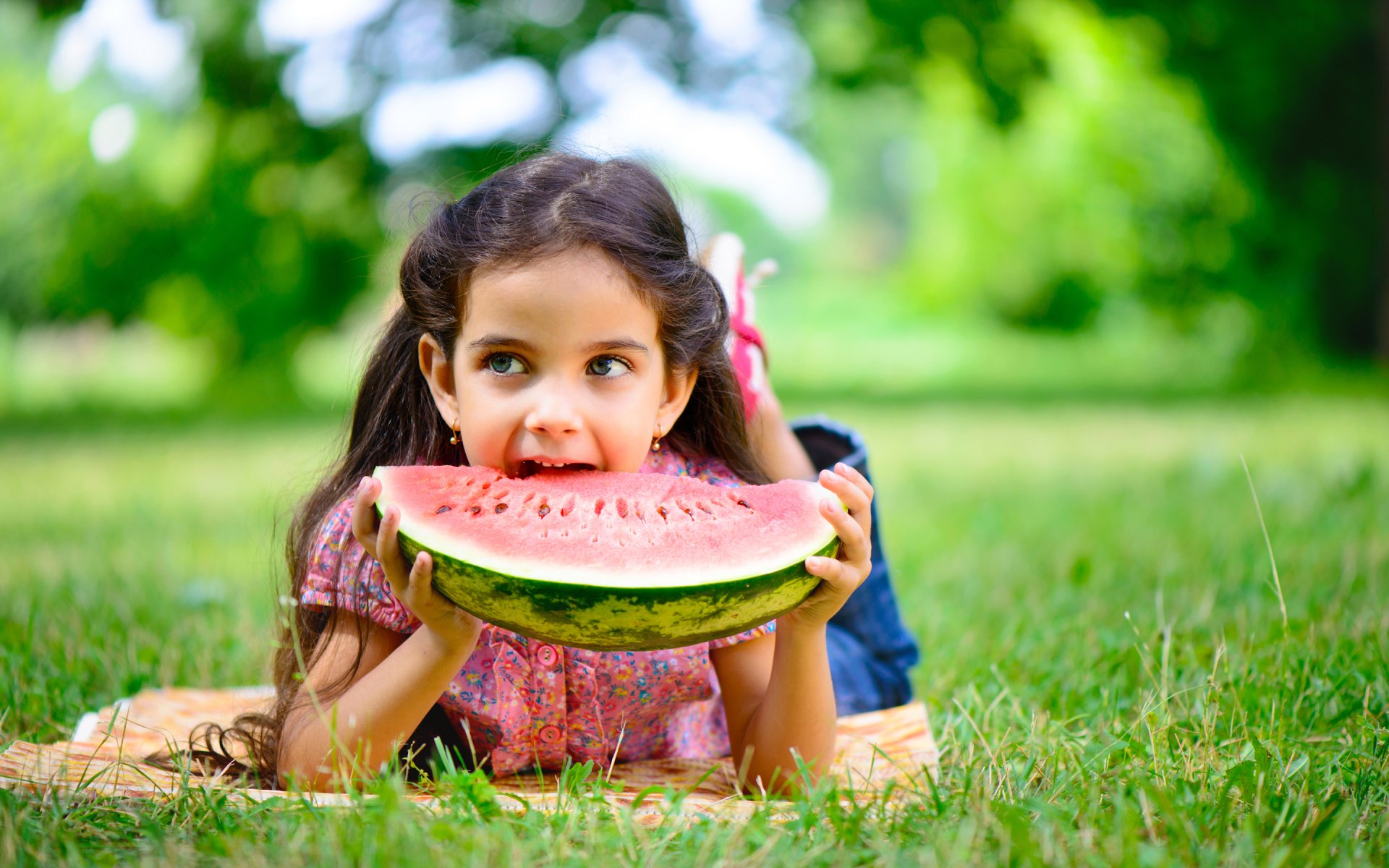 Photography — 2K Quad HD PC desktop wallpaper showing a child lying on grass in a sunlit park, smiling while biting a large watermelon slice.