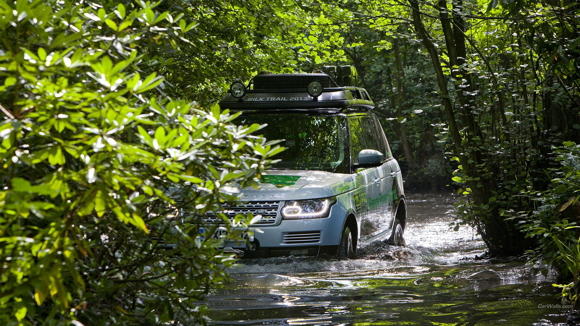HD PC desktop wallpaper: 2015 Land Rover Range Rover Hybrid fording a shallow forest stream, surrounded by lush green foliage.