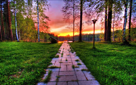 A scenic park pathway lined with lush grass and trees leads to a tranquil lake at sunset, creating a serene atmosphere in this HD desktop wallpaper.