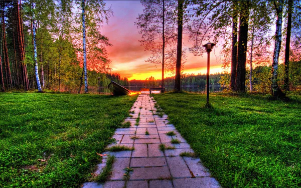 A scenic park pathway lined with lush grass and trees leads to a tranquil lake at sunset, creating a serene atmosphere in this HD desktop wallpaper.