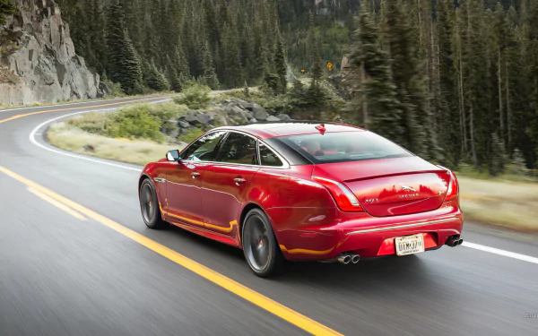 HD PC desktop wallpaper background: red Jaguar XJR vehicle driving along a winding mountain road, rear three-quarter view framed by tall pine forests.
