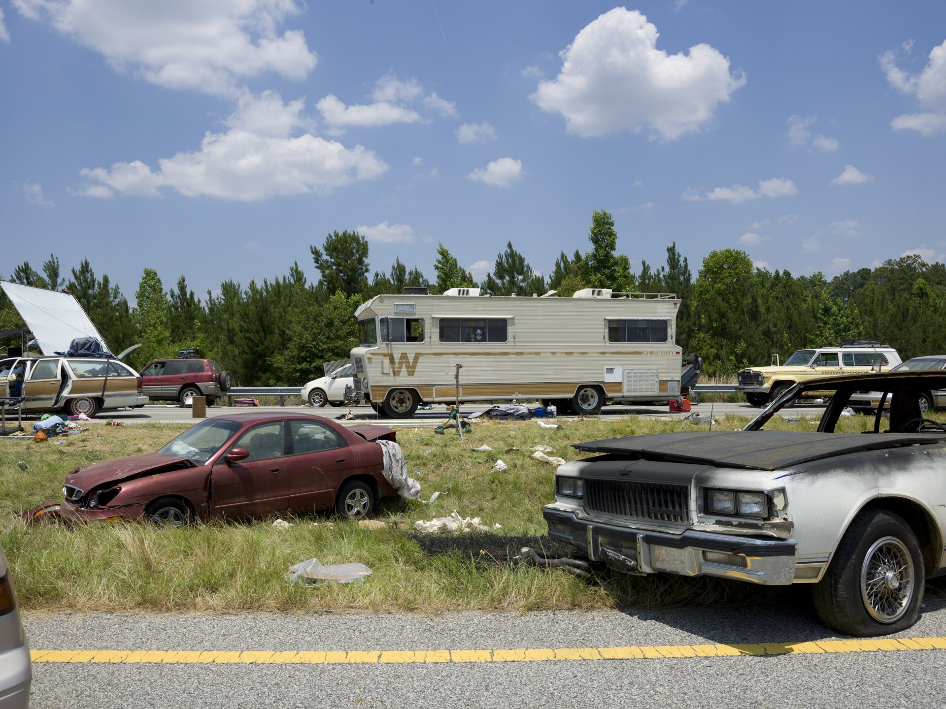 A desolate scene from The Walking Dead, featuring abandoned vehicles and an RV amidst overgrown grass under a clear blue sky, capturing the show's post-apocalyptic setting.