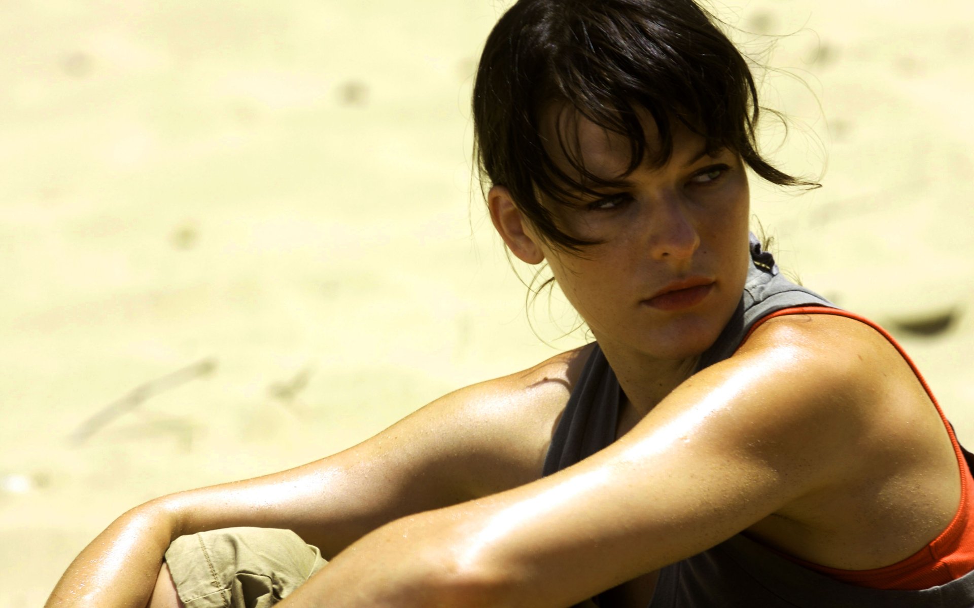HD desktop wallpaper featuring Milla Jovovich from the movie A Perfect Getaway, showing her sitting and gazing thoughtfully against a sandy background.
