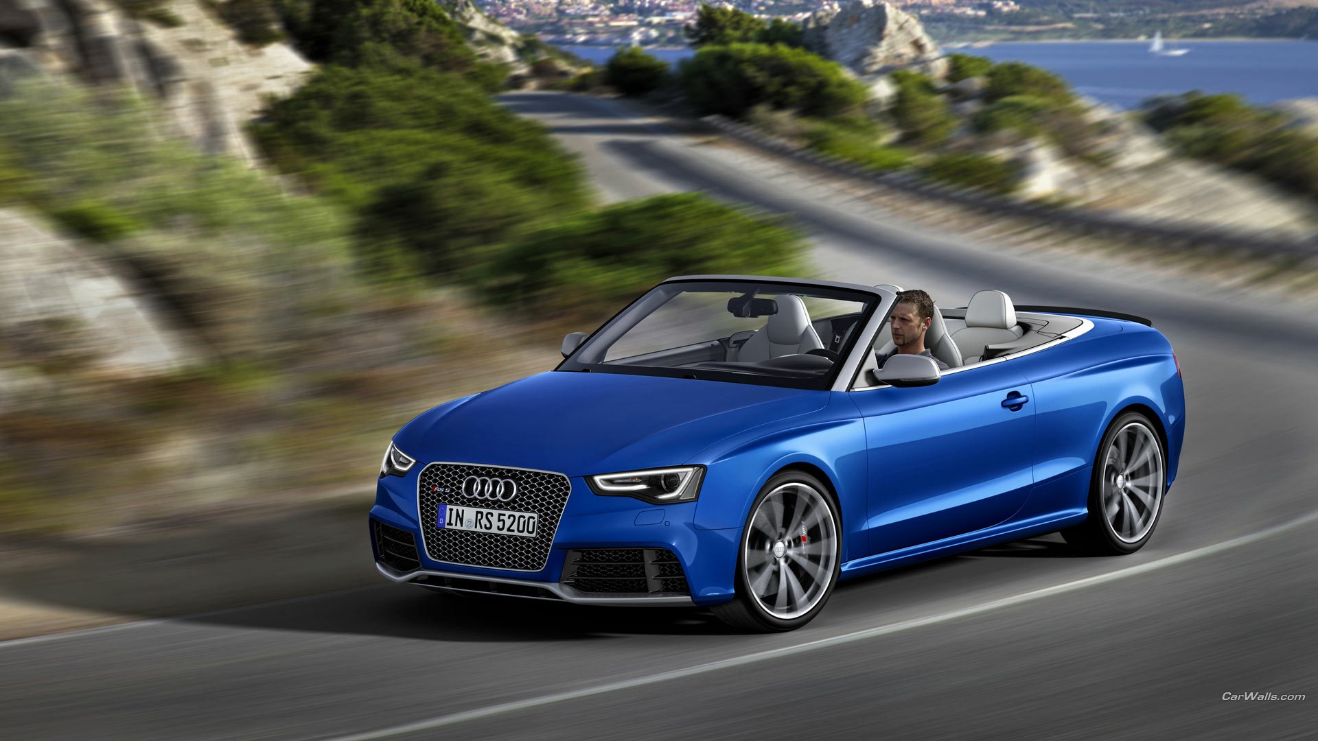 HD PC desktop wallpaper/background: blue Audi RS5 convertible vehicle speeding along a scenic coastal road, motion-blurred cliffs and ocean in the background.