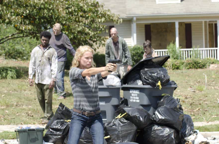 Andrea from The Walking Dead, portrayed by Laurie Holden, aims a firearm amid a zombie threat, surrounded by garbage bins and other survivors in a tense outdoor scene.