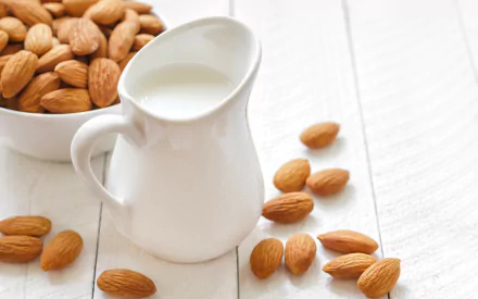 Aesthetic HD desktop wallpaper featuring a bowl of almonds alongside a small pitcher of almond milk, with scattered almonds on a light wooden surface.