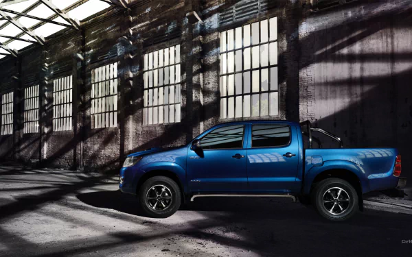 HD PC desktop wallpaper featuring a blue Toyota Hilux parked inside a large industrial building with dramatic light and shadow patterns on the walls.