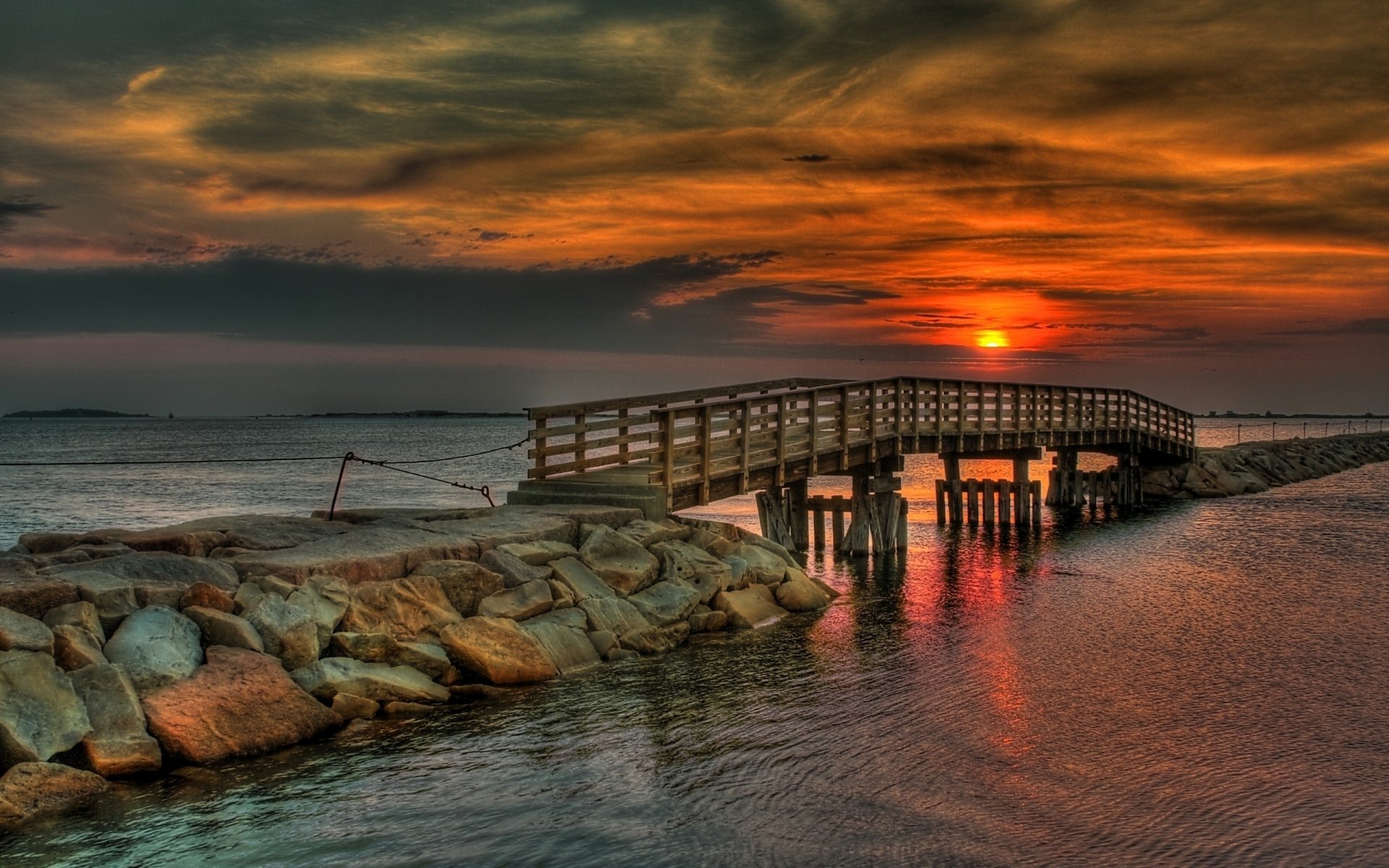 A stunning HD desktop wallpaper featuring a wooden bridge over tranquil waters at sunset, surrounded by a rocky shoreline and dramatic clouds.