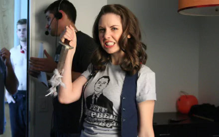 A dynamic HD wallpaper featuring Alison Brie, showcasing her playful energy as she raises her fist in a humorous pose while wearing a graphic tee and a vest.