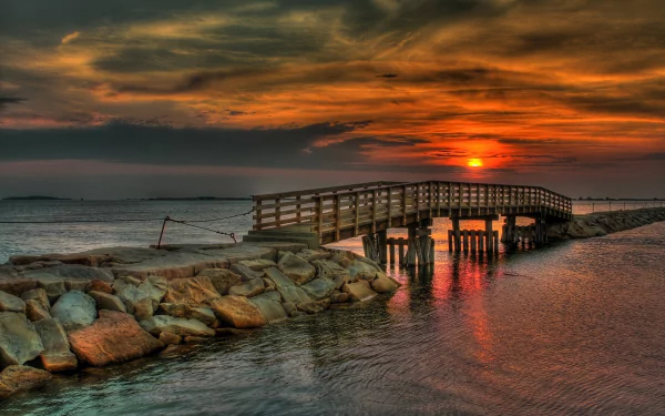 A stunning HD desktop wallpaper featuring a wooden bridge over tranquil waters at sunset, surrounded by a rocky shoreline and dramatic clouds.